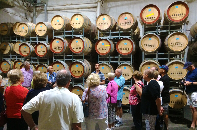 Griffith Tours Home Page picture of a group of people visiting a winery with a stack of large wine barrels in the background.